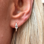 A close-up of a person's ear with light blonde hair, wearing the Pavé Huggie with Emerald Cut Diamonds, a gold hoop earring featuring emerald cut diamonds and rectangular clear gemstones.
