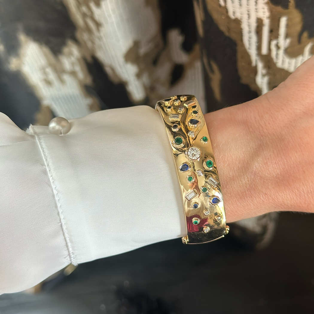 A close-up of a wrist wearing the Bezel Set Mixed Shape Emerald, Diamond, and Sapphire Cuff in Yellow Gold, paired with a white shirt featuring a pearl button and a patterned outfit.