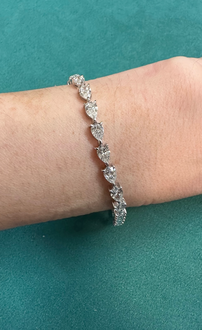 The Chasing Pears Diamond Tennis Bracelet, featuring heart-shaped clear stones set in silver, is worn on a wrist and displayed against a teal background.