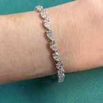 The Chasing Pears Diamond Tennis Bracelet, featuring heart-shaped clear stones set in silver, is worn on a wrist and displayed against a teal background.