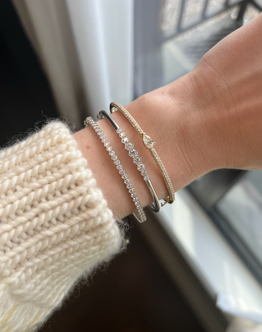 A wrist adorned with the Hexagon Bezel Set Diamond Bangle in 18k gold and two silver gemstone bracelets, styled with a cream knit sweater, captured indoors by a window.
