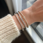 A wrist adorned with the Hexagon Bezel Set Diamond Bangle in 18k gold and two silver gemstone bracelets, styled with a cream knit sweater, captured indoors by a window.