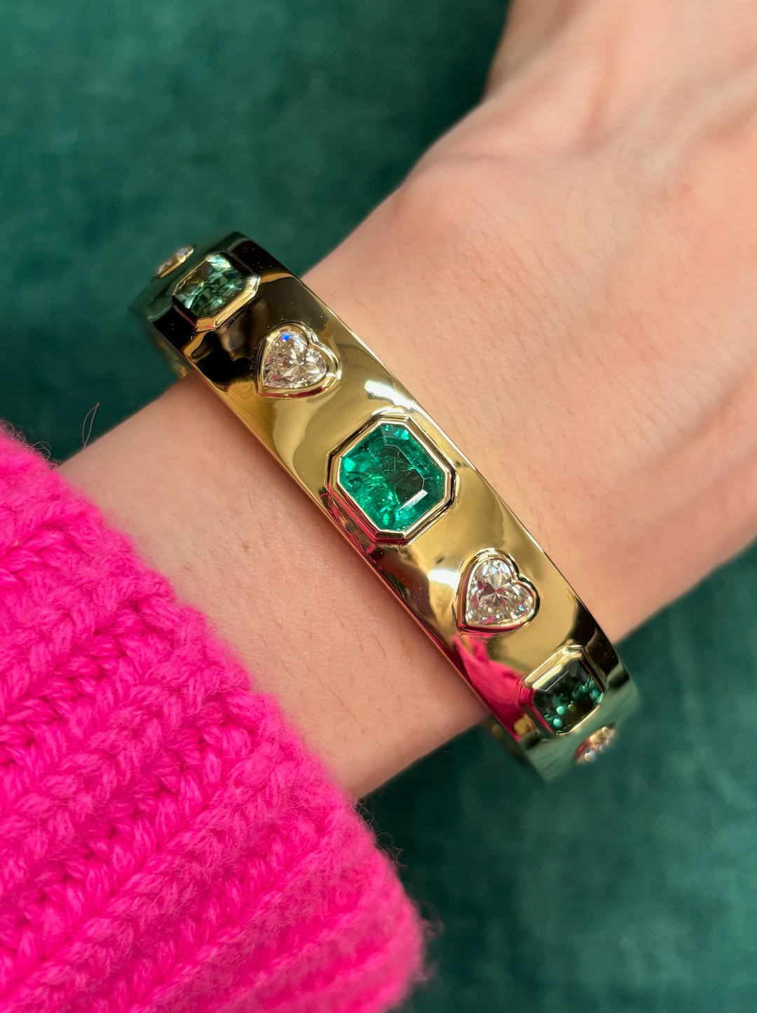 The Bezel Set Mixed Shape Diamond and Emerald Bangle in Yellow Gold shines on a wrist adorned with a bright pink, ribbed knit sweater, its emeralds and diamonds glowing against a lush green backdrop.
