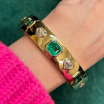 The Bezel Set Mixed Shape Diamond and Emerald Bangle in Yellow Gold shines on a wrist adorned with a bright pink, ribbed knit sweater, its emeralds and diamonds glowing against a lush green backdrop.