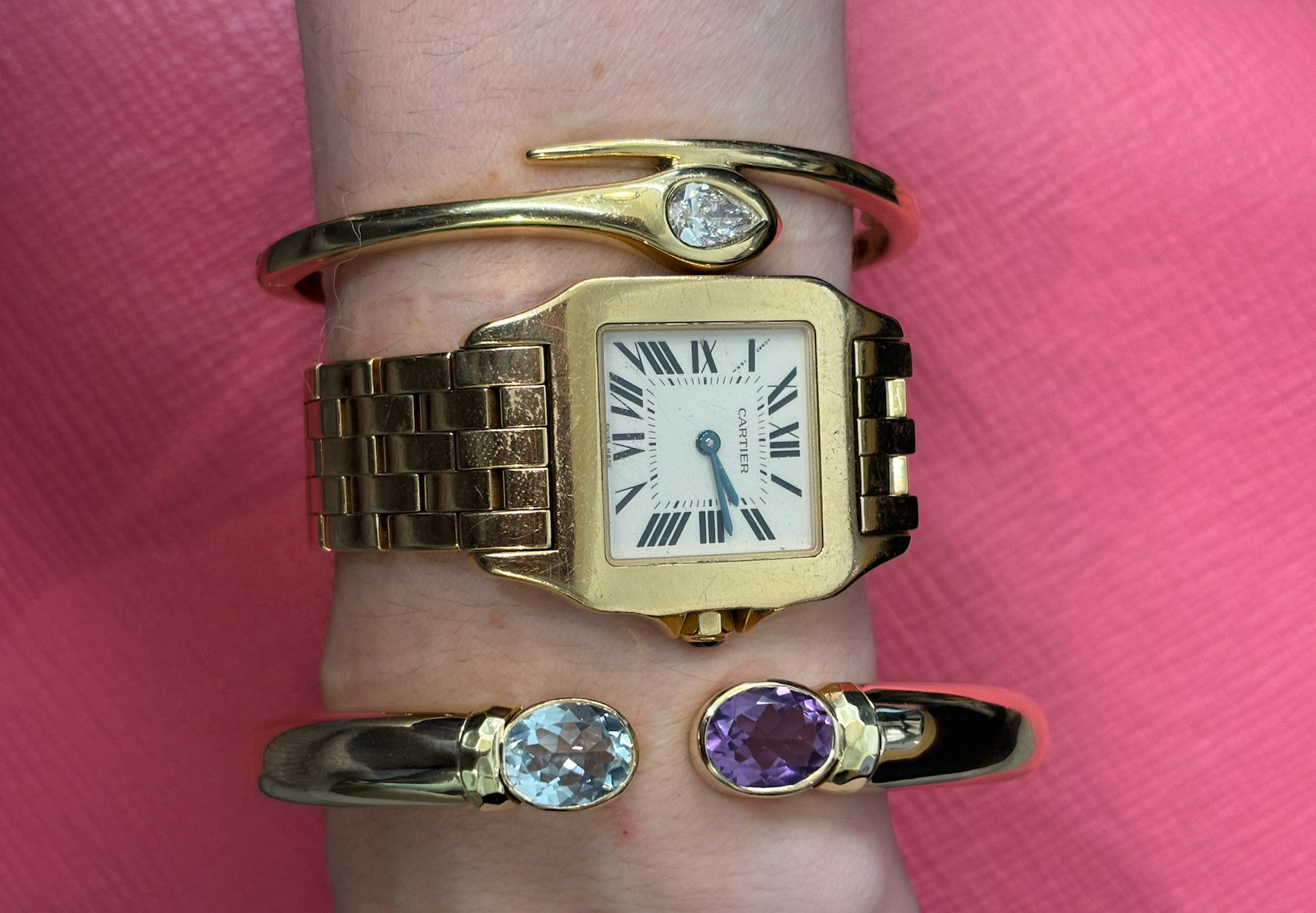 A wrist sporting an 18K yellow gold square watch with Roman numerals, paired with a gold nail-design bracelet and the Bezel Set Open Space bangle featuring oval aquamarine and amethyst, set against a pink background.