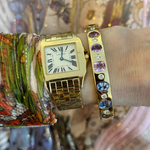 A wrist with a gold Cartier square-face watch with Roman numerals and the Burnish Set Multicolor, Mixed Shape, Diamond and Sapphire Bangle in Yellow Gold, set against colorful, patterned fabric.