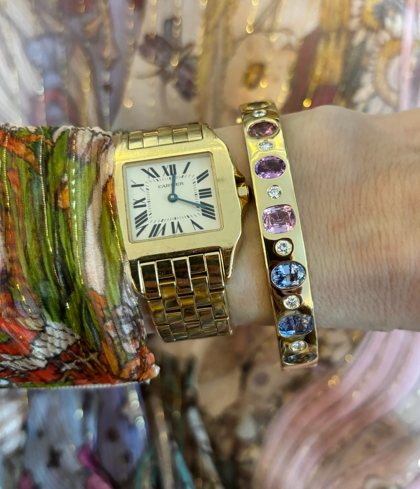 A wrist with a gold Cartier square-face watch with Roman numerals and the Burnish Set Multicolor, Mixed Shape, Diamond and Sapphire Bangle in Yellow Gold, set against colorful, patterned fabric.