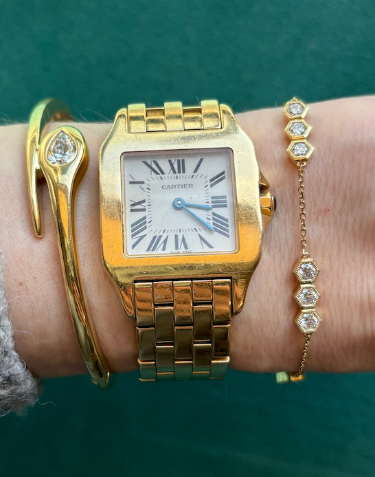 A wrist adorned with a gold Cartier watch with a square face and Roman numerals, an 18k gold nail-shaped bracelet with a diamond, and the Serpentine Station Bracelet with Hexagon Diamond Trios.