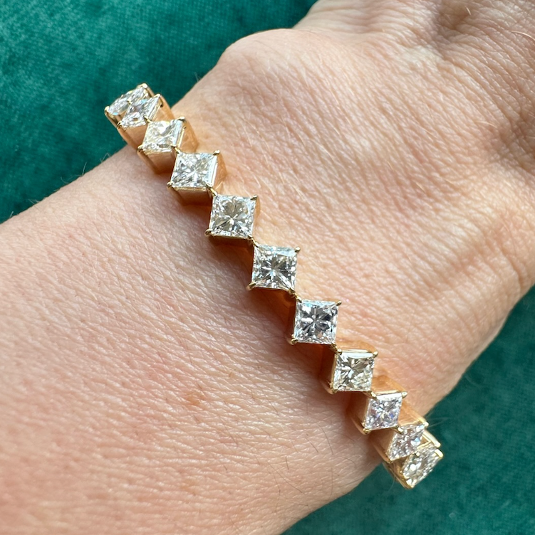 The Diagonally Set Square Diamond Bangle in Yellow Gold features square-cut diamonds in 18K yellow gold, gleaming on the wrist against a teal backdrop.