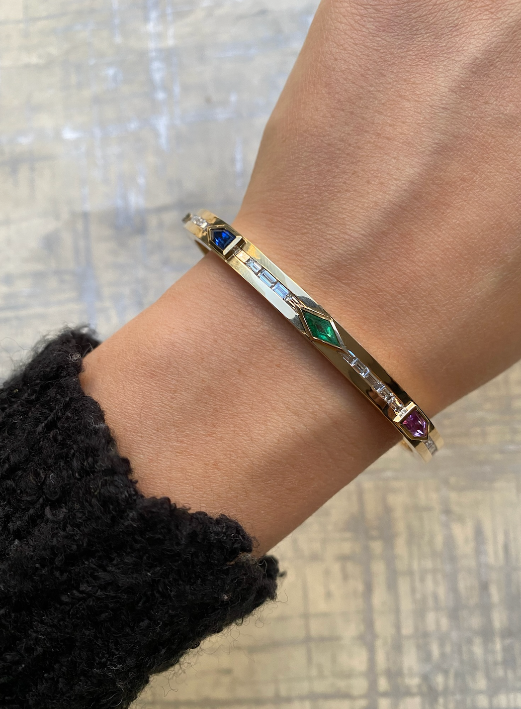 A close-up of a wrist adorned with the Bezel Set Mixed Shape Sapphire, Emerald, and Baguette Diamond Bangle. The vibrant gems and geometric design pop against a textured, light-colored background.