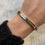 A close-up of a wrist adorned with the Bezel Set Mixed Shape Sapphire, Emerald, and Baguette Diamond Bangle. The vibrant gems and geometric design pop against a textured, light-colored background.