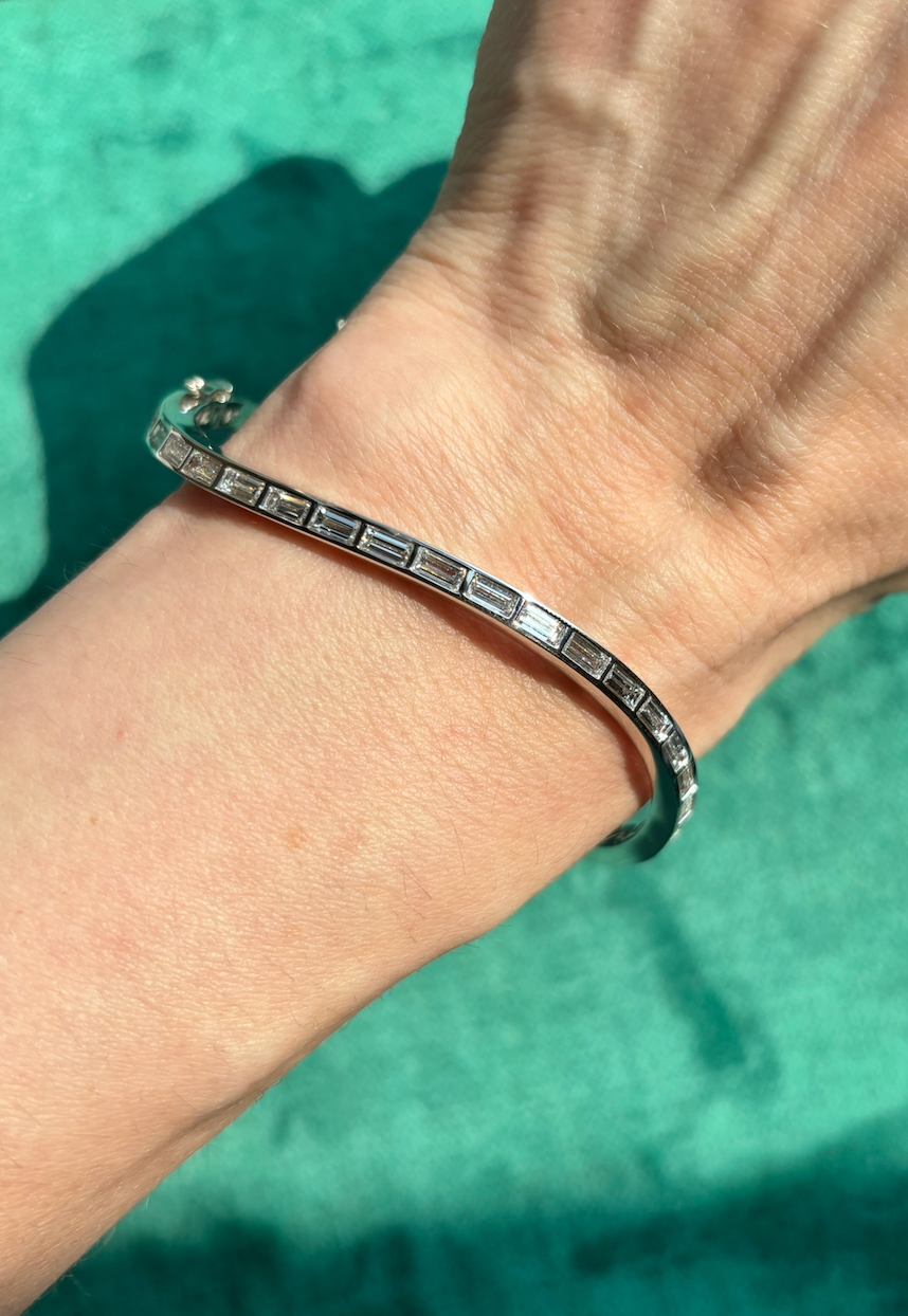 The Burnish Set Baguette Diamond Wave Bangle in White Gold adorns a wrist, its sleek design shining in bright sunlight against a textured green backdrop.