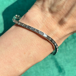 The Baguette Diamond Wave Bangle in White Gold, featuring sleek rectangular links and minimalist design, sparkles elegantly on the wrist against a green textured background.