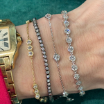 A wrist wearing a gold Cartier watch and four delicate diamond bracelets, including the Black Rhodium Hexagon Bezel Set Diamond Tennis Bracelet and others in yellow gold, rose gold, white gold, and black rhodium, set against a green background.