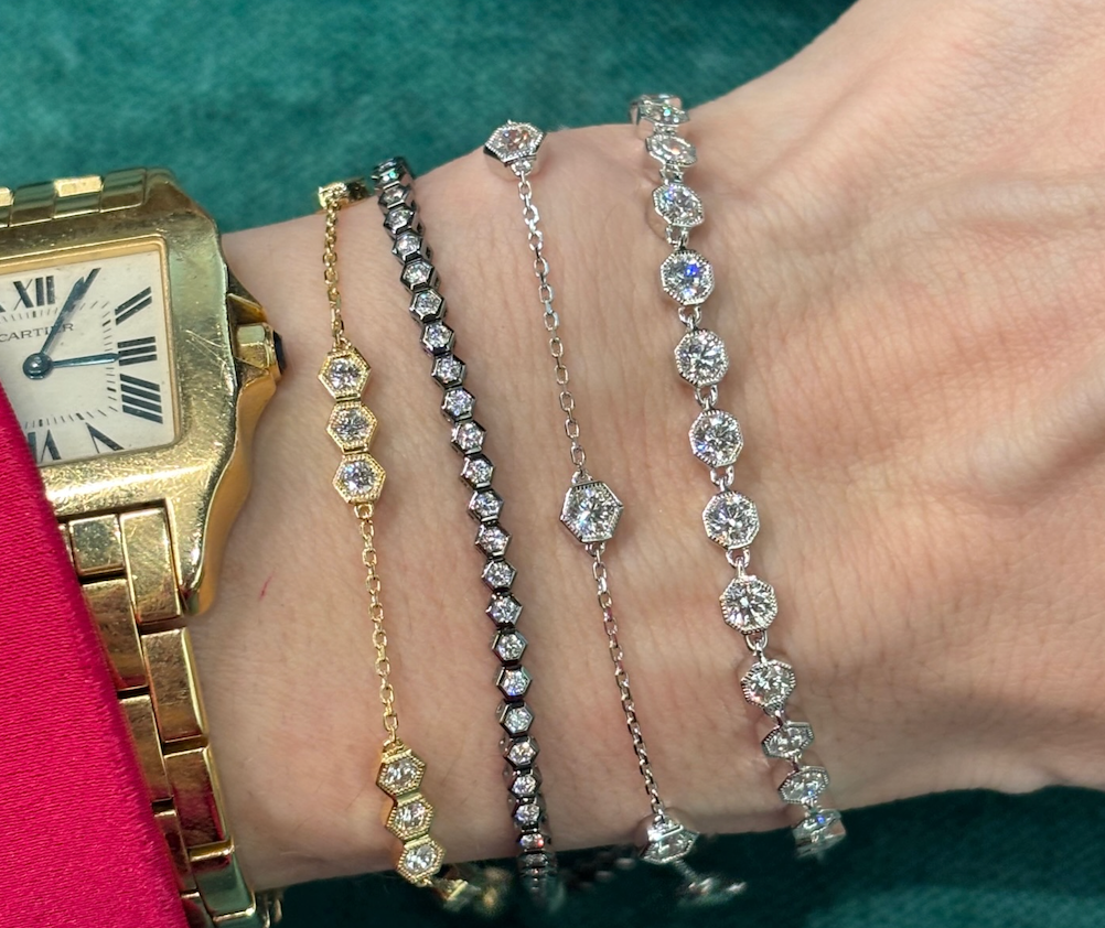 A wrist wearing a gold Cartier watch and four delicate diamond bracelets, including the Black Rhodium Hexagon Bezel Set Diamond Tennis Bracelet and others in yellow gold, rose gold, white gold, and black rhodium, set against a green background.