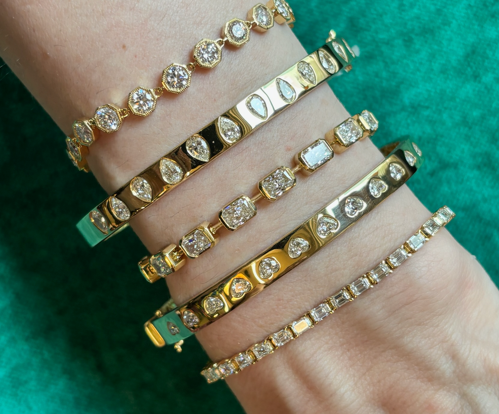 A wrist wears five East-West Half Bezel Set Radiant Cut Diamond Tennis Bracelets in yellow gold, showcased against a green background.