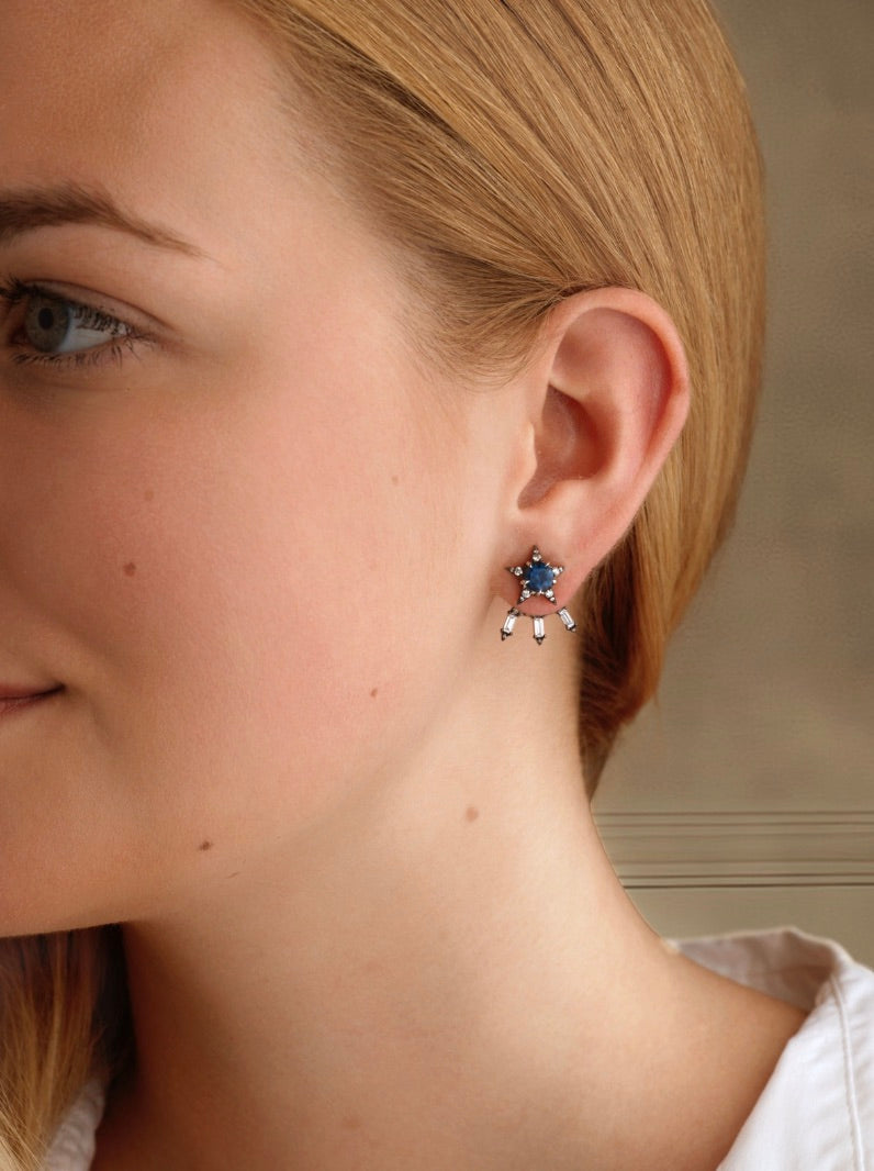 A close-up of a woman’s ear wearing the America250 Baguette Diamond Ear Jackets in Black Rhodium, featuring blue and red stones with dangling rectangular accents, handcrafted in NYC.
