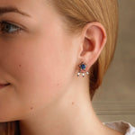 A close-up of a woman’s ear wearing the America250 Baguette Diamond Ear Jackets in Black Rhodium, featuring blue and red stones with dangling rectangular accents, handcrafted in NYC.