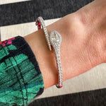 A person wearing the Pavé Serpent Diamond Bangle—18K white gold with pear-shaped diamond accents—dressed in a green and black floral sleeve. The background shows a black and white striped surface.