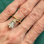 A close-up of a hand wearing the Shield Diamond Perch Engagement Ring in yellow gold, featuring a large uniquely cut clear gemstone, against a green fabric background.