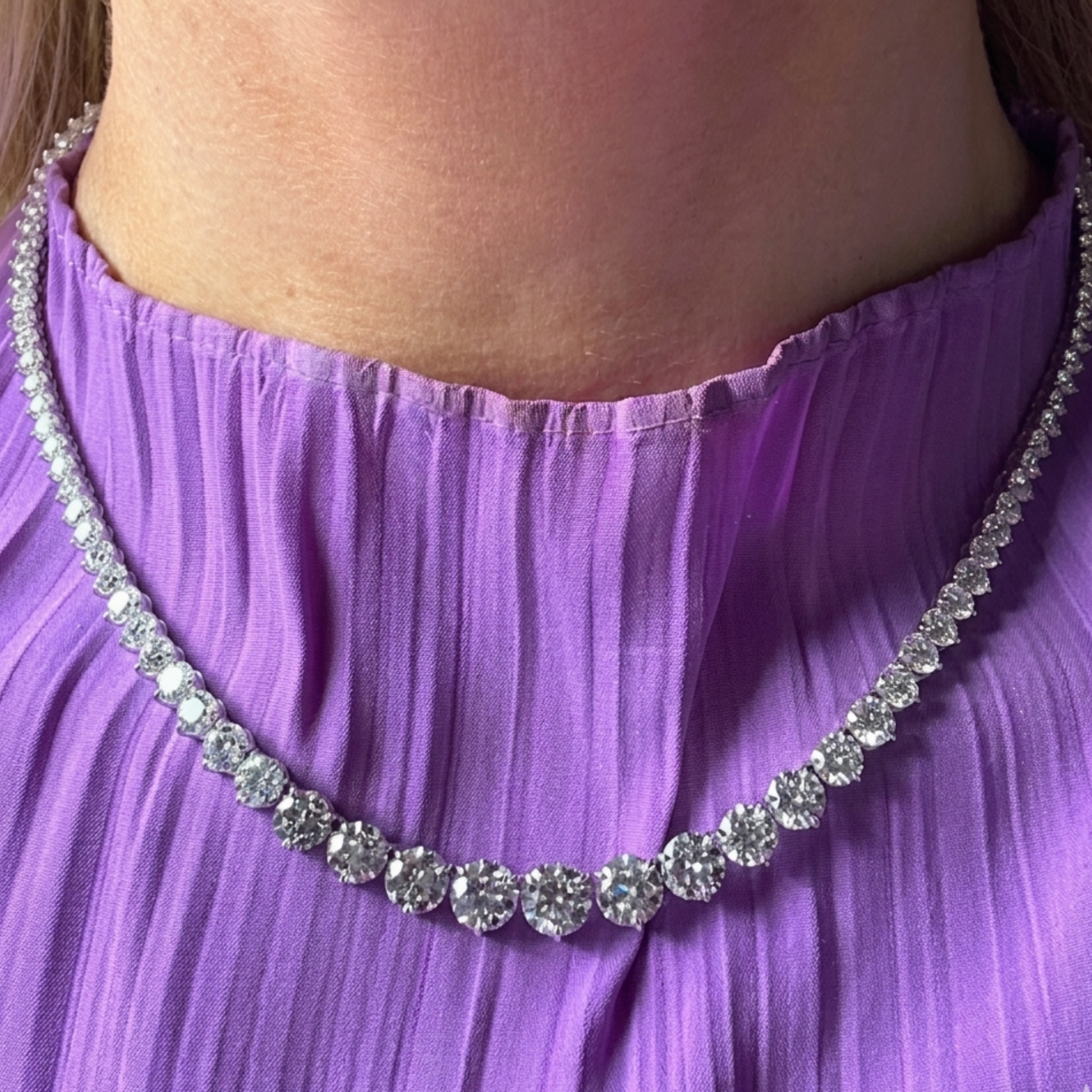A person wearing a pleated, purple top and the Small Graduated Round Diamond Tennis Necklace featuring sparkling round diamonds set in 18k gold, with larger stones at the center and smaller ones toward the clasp.