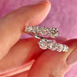 A hand holds the Graduated Round Diamond Wraparound Ring in white gold, featuring a row of sparkling round diamonds, set against a soft pink background.