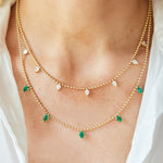 A close-up of a person wearing two layered necklaces, including the Spike Necklace in Yellow Gold with Pear Emeralds and a station necklace with crystal drops, set against light skin and a white shirt.