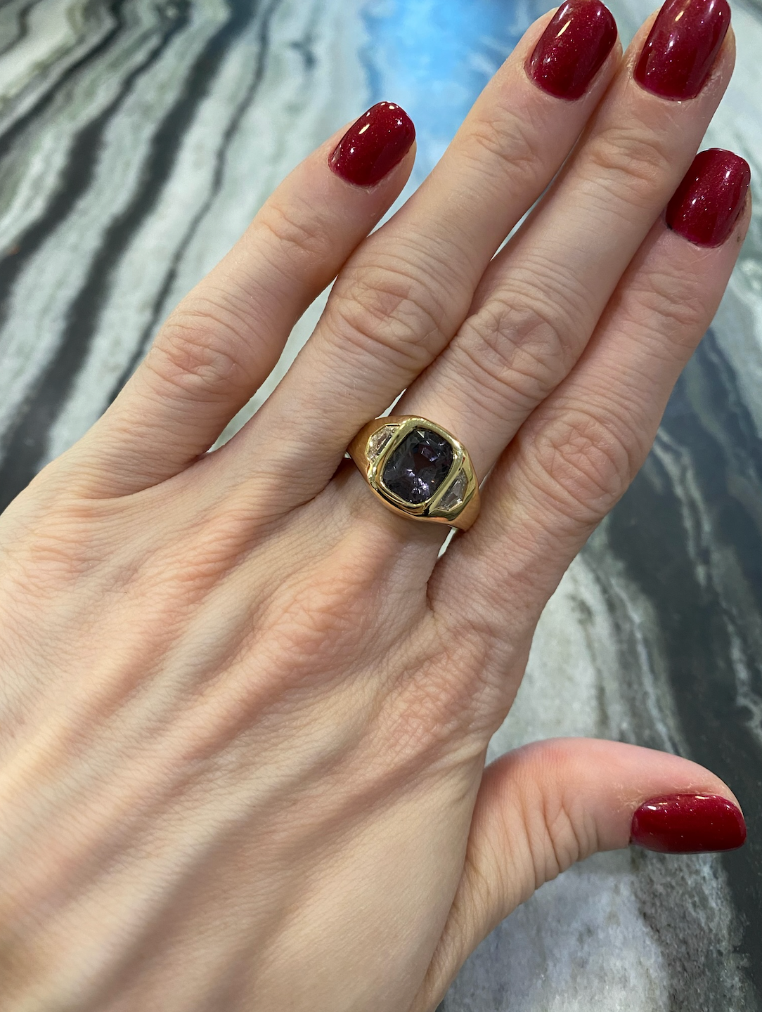 A hand with red-painted nails showcases the Bespoke Cushion Cut Spinel Signet Ring in Yellow Gold with Epaulette Diamond Side Stones, set against a wavy-patterned gray and black background.