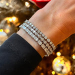 A close-up of a wrist wearing three diamond bracelets, featuring the Round Brilliant Diamond Tennis Bracelet in White Gold, set against a warm, festive background with golden lights and holiday decor.