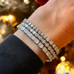 A close-up of a wrist wearing the Large Hexagon Bezel Set Diamond Tennis Bracelet in 18K gold, sparkling against a blurred, warmly lit background. The person wears a black sleeve.