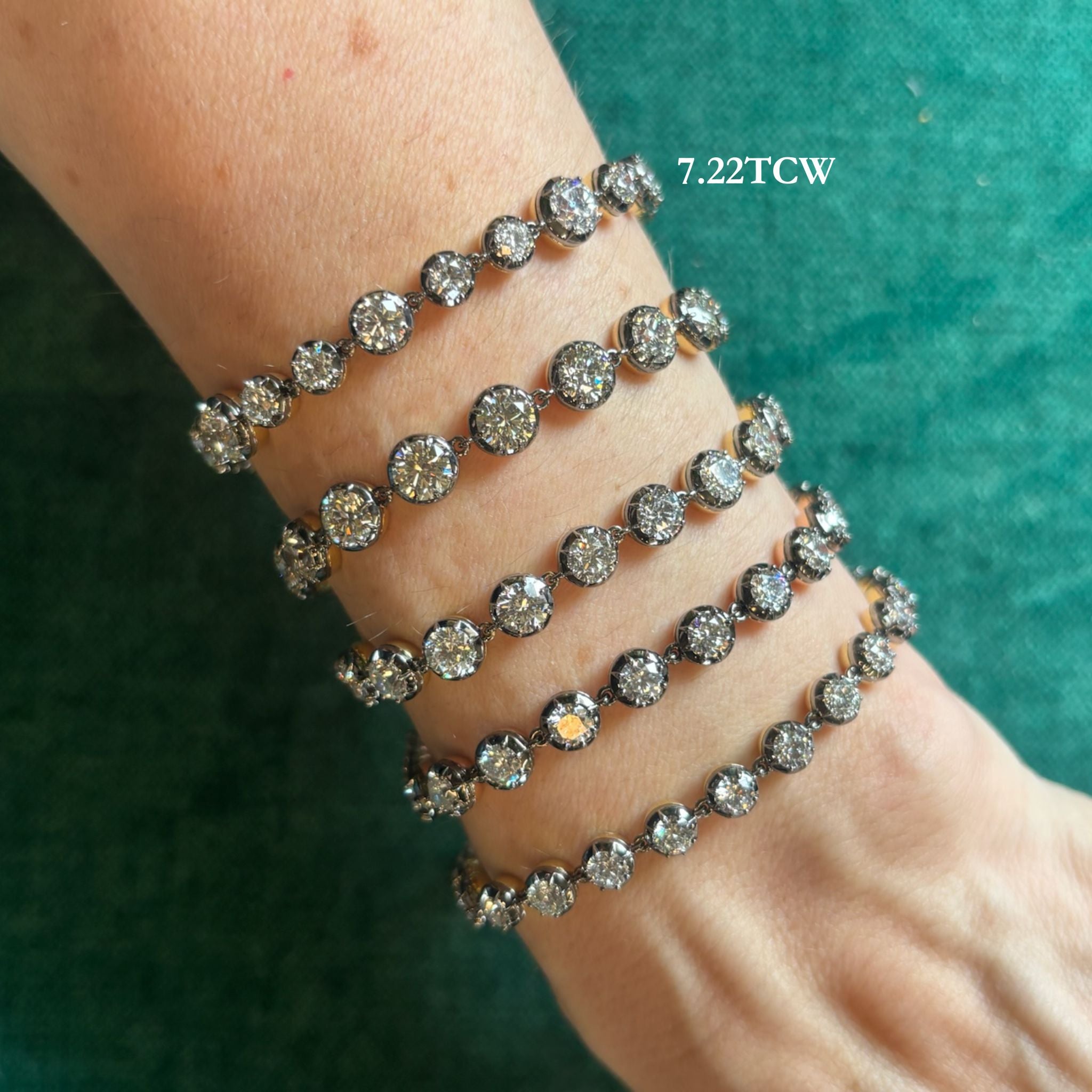 A wrist is adorned with five dazzling Collet Set Round Diamond Bracelets in black rhodium, their round diamonds sparkling beautifully against a green textured backdrop. Text on the image reads 7.22 TCW.