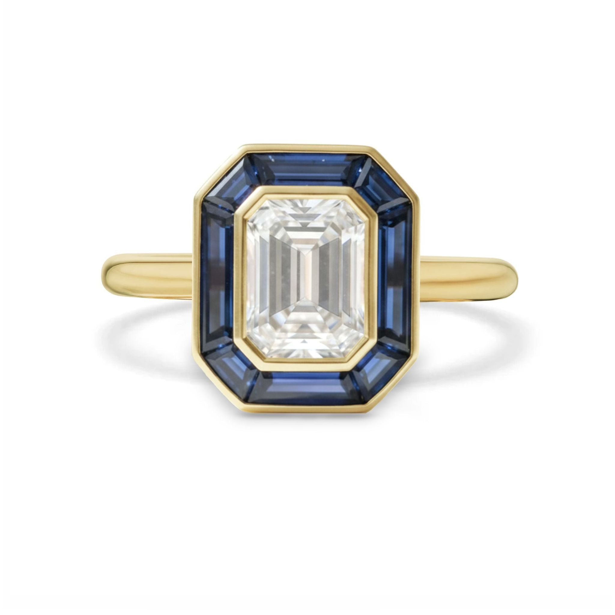The Mosaic Bezel Set Emerald Cut Diamond Engagement Ring features a central emerald-cut diamond with a trapezoid blue sapphire halo, all set in yellow gold.
