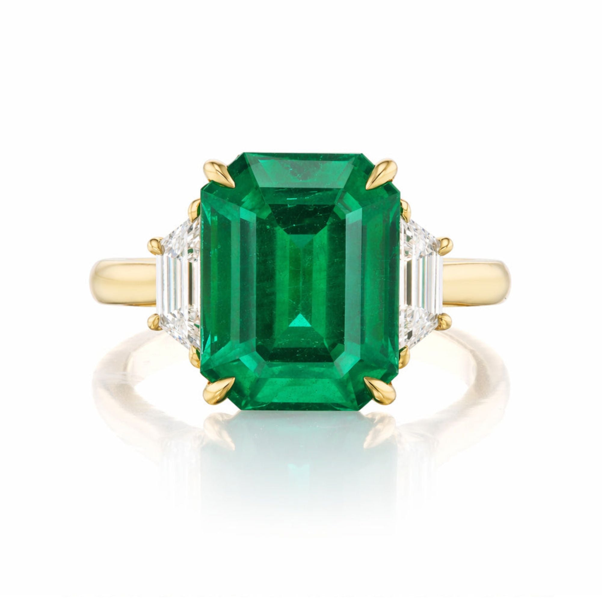 The Emerald Cut Emerald Engagement Ring features a central emerald with trapezoid baguette diamond side stones, all set in yellow gold and displayed against a white background.