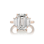 The Emerald Cut Diamond Engagement Ring with Trapezoid Side Stones features a large emerald-cut diamond at the center, flanked by two smaller trapezoid diamonds on each side, and is shown in elegant rose gold against a white background.
