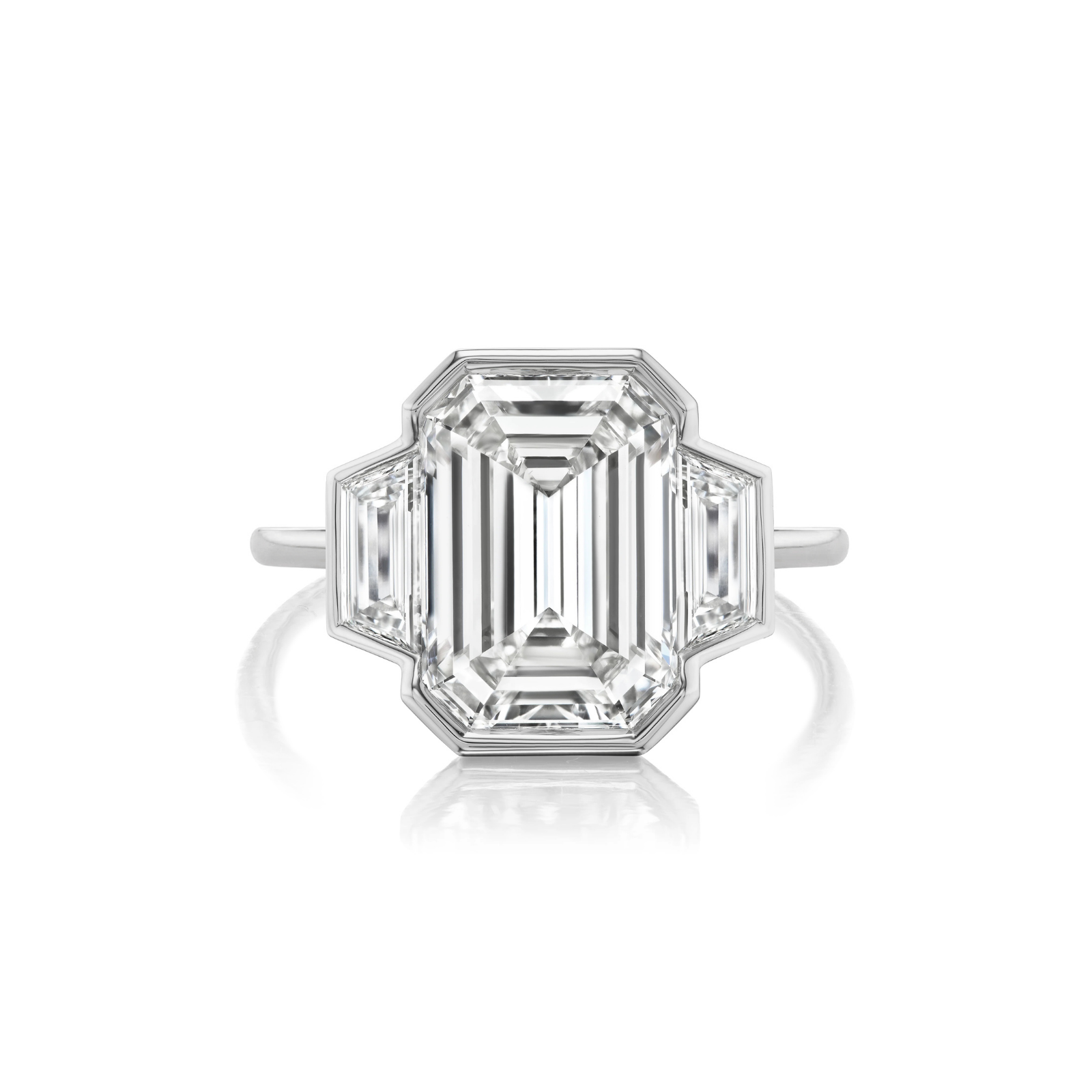 Introducing our Half Bezel Set Three Stone Engagement Ring, showcasing a stunning emerald-cut diamond at the center, flanked by trapezoid side stones and elegantly set on a slim platinum band.