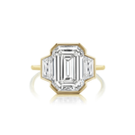 A Half Bezel Set Three Stone Engagement Ring with an emerald cut diamond center and trapezoid side stones is displayed on a white background, featuring a large, step-cut center stone in a geometric, octagonal setting.