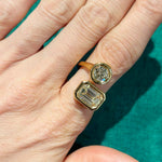 A hand wearing the Toi et Moi Diamond Wraparound Ring, bezel set in 18k yellow gold with round and emerald cut diamonds side by side, is displayed against a green background, highlighting the timeless beauty of diamond jewelry.