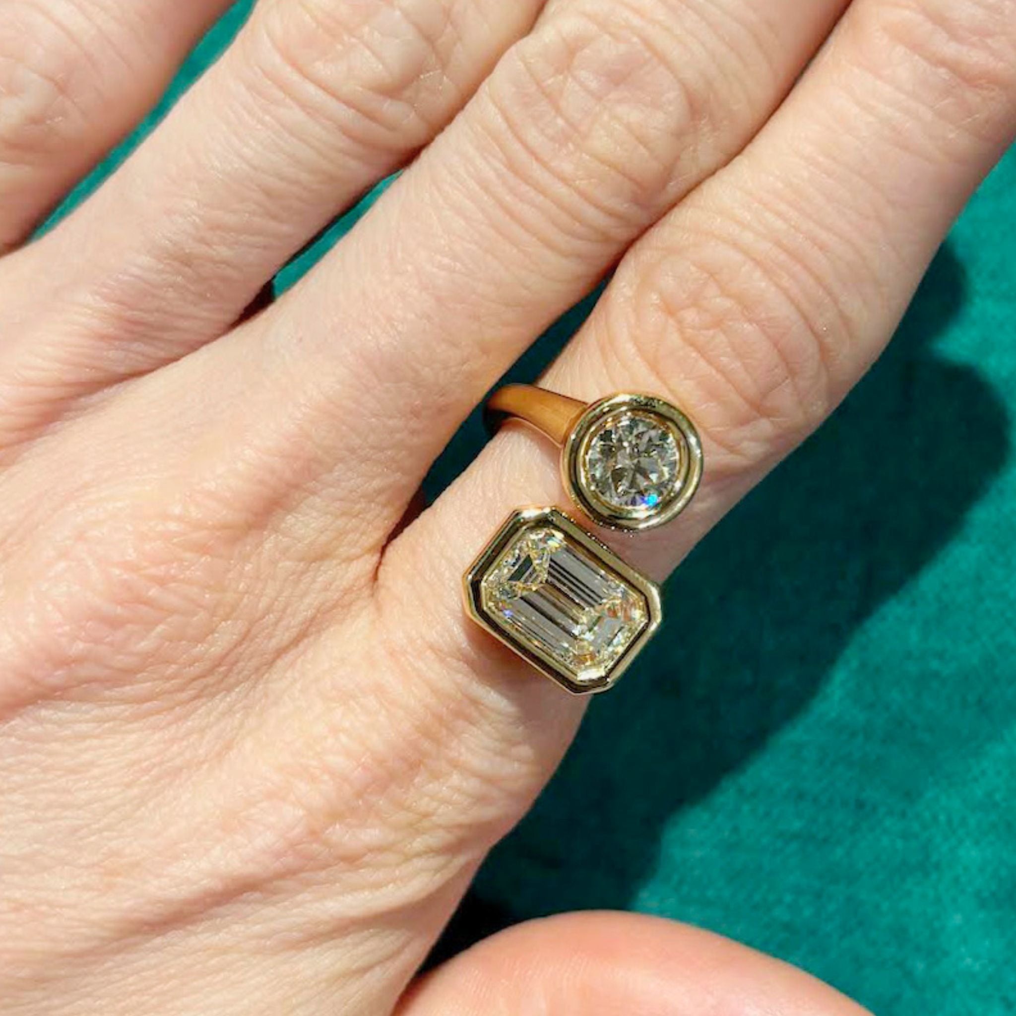 A hand wearing the Toi et Moi Diamond Wraparound Ring, bezel set in 18k yellow gold with round and emerald cut diamonds side by side, is displayed against a green background, highlighting the timeless beauty of diamond jewelry.