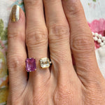 A hand with gold-painted nails wears the Toi et Moi Pavé Ring, featuring a square emerald cut diamond and a pink emerald cut sapphire set side by side in 18k yellow gold. The background includes a floral fabric pattern.