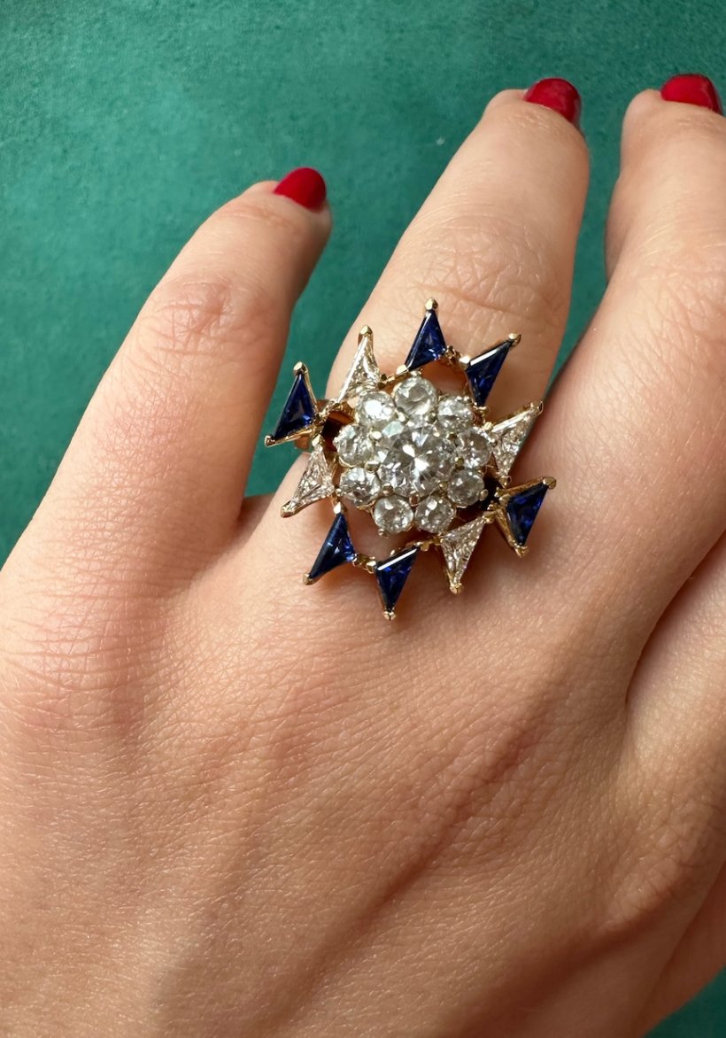 A hand with red polish showcases the Bespoke Triangle Diamond and Sapphire Ring Jacket in Yellow Gold, featuring a central cluster of clear gems and alternating sapphire and white triangular stones, set against a green background.