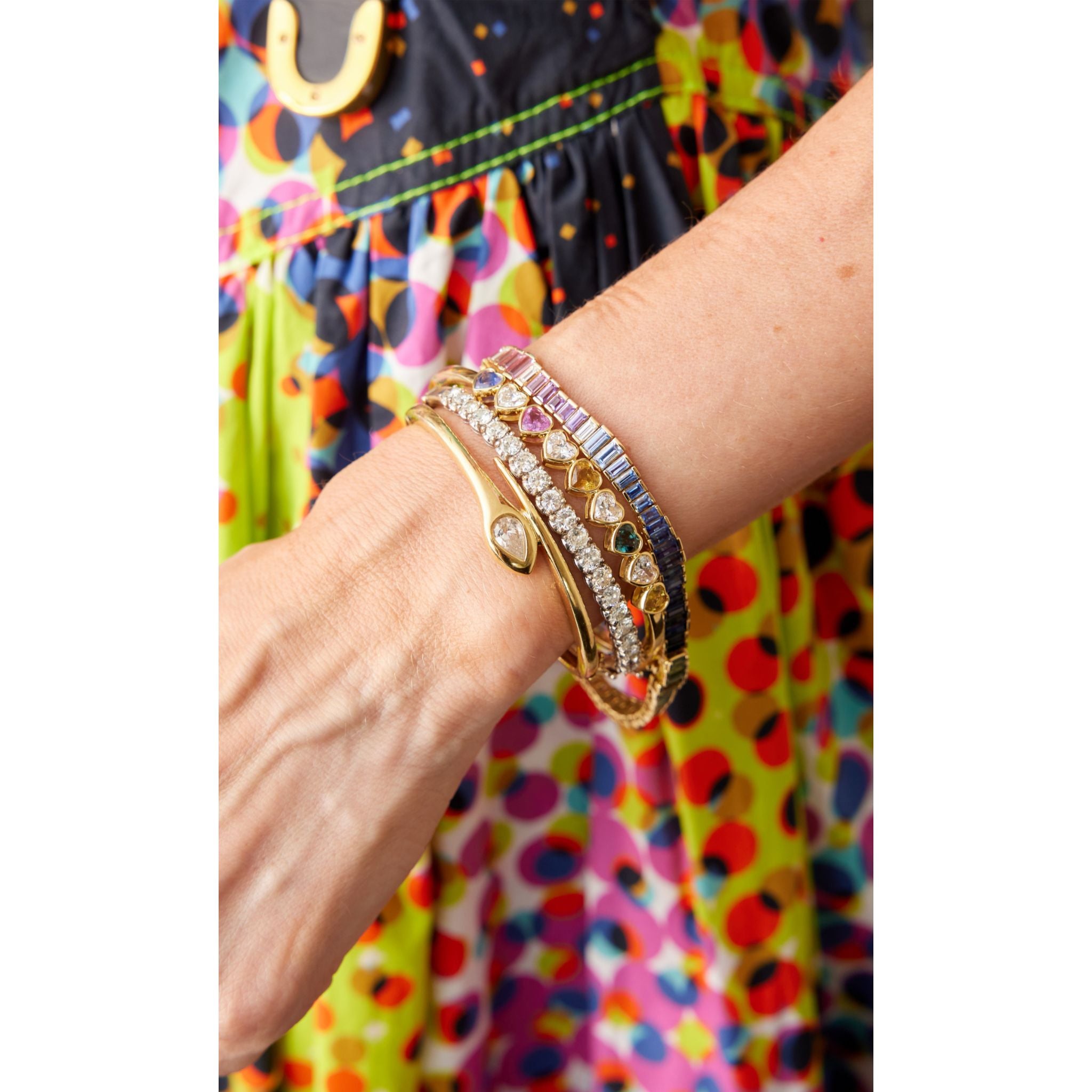 A close-up of a wrist adorned with multiple Chroma Heart Beats Bezel Set Heart Shape Sapphire and Diamond Bangles in yellow gold, set against a colorful polka-dotted dress background.