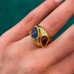 A hand wearing a Bulgari Pink Tourmaline and Sapphire Ring, circa 1980, featuring two marquise-shaped gemstones—one blue and one deep red—set side by side on a green background.