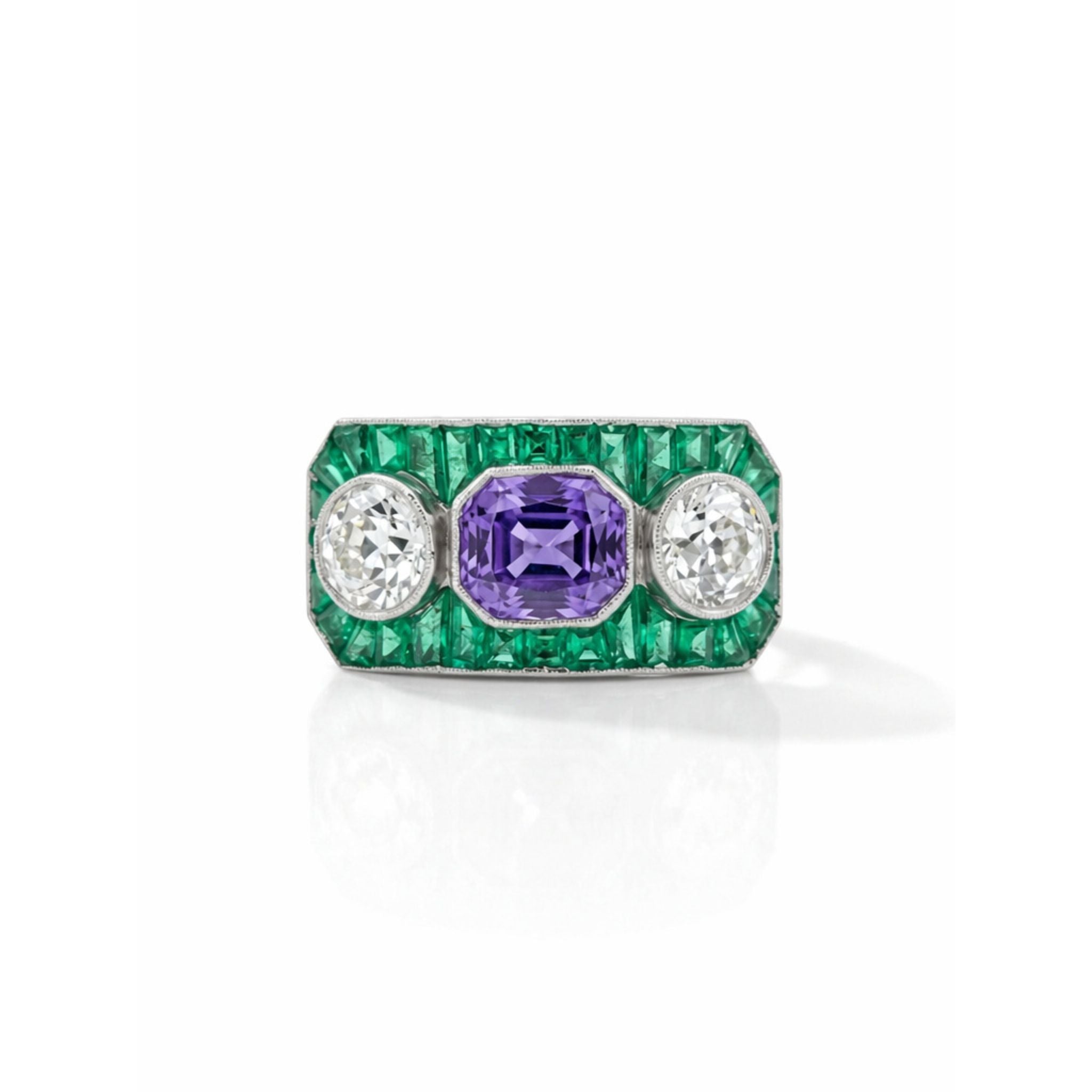The Sapphire, Emerald and Diamond Ring features a rectangular emerald-cut purple sapphire flanked by two round diamonds and bordered by green emeralds, evoking 1930s platinum elegance against a white background.