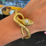 The Antique Gold, Diamond and Emerald Bracelet features a coiled snake design with a jeweled head adorned by a clear diamond and vibrant emerald-green eyes, elegantly wrapped around the wearer’s wrist.