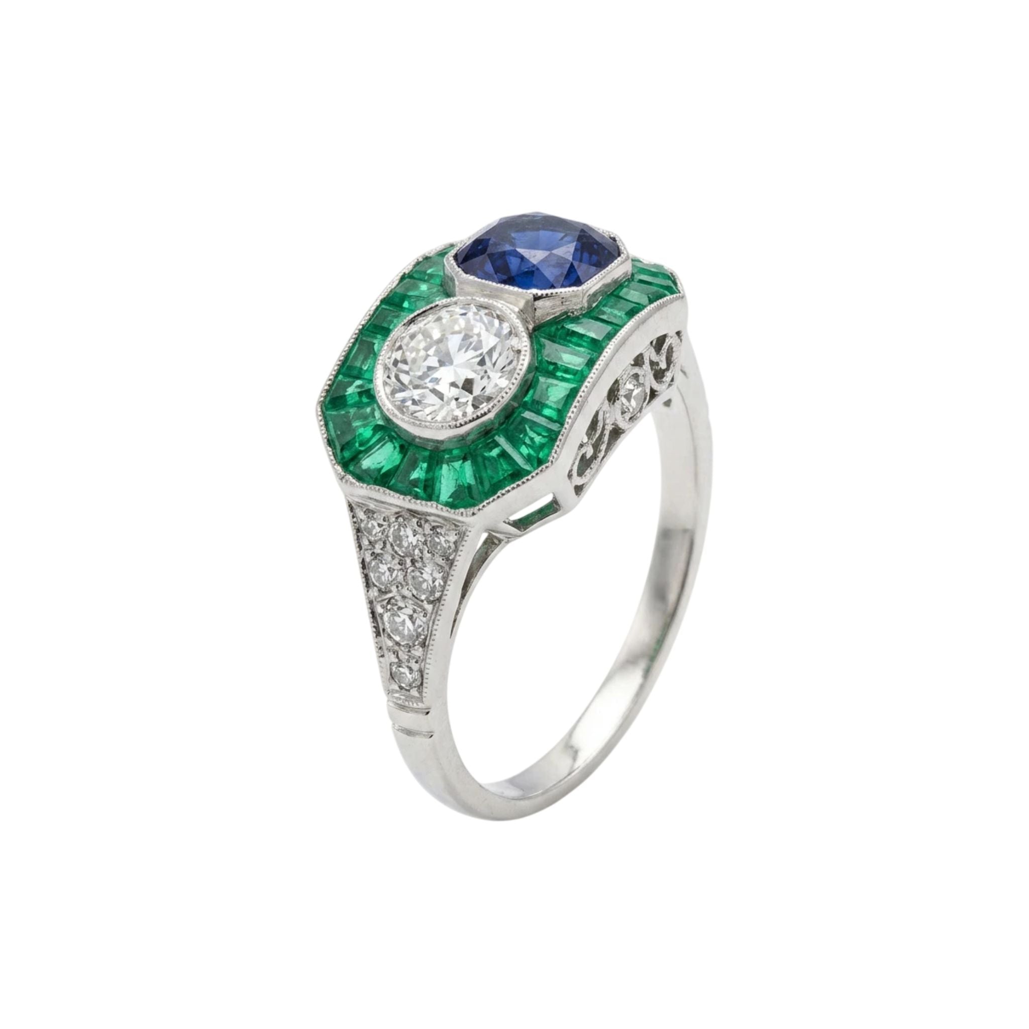 The Sapphire, Emerald and Diamond Ring features a round white diamond with a blue sapphire, encircled by green emerald-cut stones and small diamonds on a silver band inspired by 1930s platinum styles.