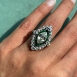 A hand displays the Art Deco Emerald Diamond Ring, featuring a large marquise diamond surrounded by smaller diamonds and emerald accents, against a green background.