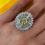 A hand wears the Mid-Century Fancy Intense Yellow Diamond and Diamond Ring, showcasing a central VVS2 Fancy Intense Yellow diamond framed by layered sparkling diamonds in a floral design, with an accompanying GIA report.