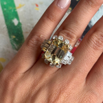 A hand with light pink nail polish displays the Yellow Sapphire and Diamond Ring, featuring an emerald-cut yellow sapphire center stone surrounded by sparkling diamonds in various cuts, set on a white gold or silver band.