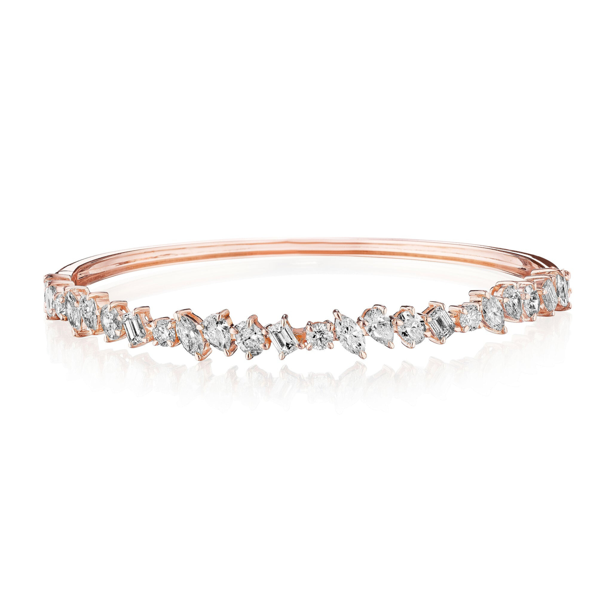 The Marching Mixed Shape Diamond Bangle, crafted in 18K rose gold with clear gemstones and emerald cut diamonds in an irregular pattern, showcases a sparkling, elegant design against a white background.