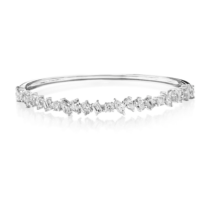 The Marching Mixed Shape Diamond Bangle in white gold showcases emerald-cut and mixed shape diamonds clustered elegantly along the front, displayed on a reflective white surface.